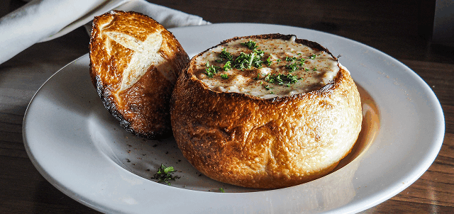 A bread bowl filled with creamy soup, garnished with herbs, sits on a white plate with its lid on the side.