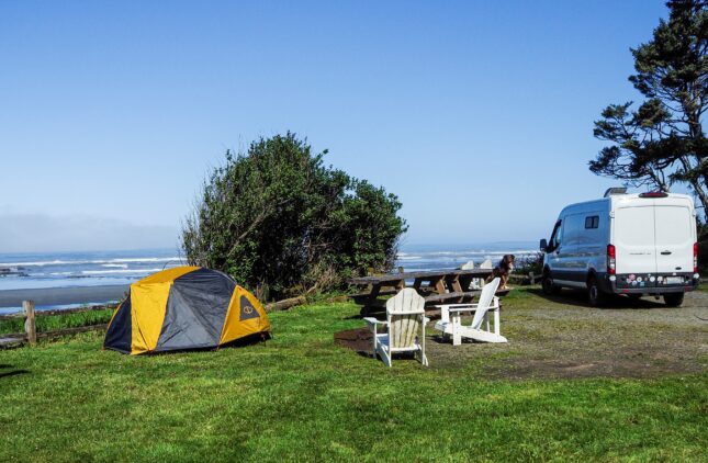 A yellow tent, white van, and chairs set up at a grassy campsite on Olympic Peninsula under a clear blue sky.