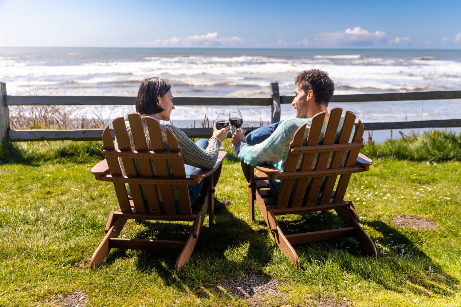 A couple relaxes in wooden chairs on Olympic Peninsula, toasting with wine under a sunny sky.