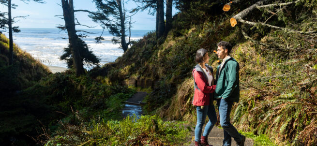 A couple stands on a forest trail near the coast on Olympic Peninsula, smiling at each other under tall trees.