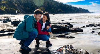 A couple kneels on a rocky beach at Olympic Peninsula, smiling and looking at sea life by the shoreline on a sunny day.