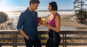 A man and woman smiling and holding drinks on a sunny beachside wooden deck at Olympic Peninsula.