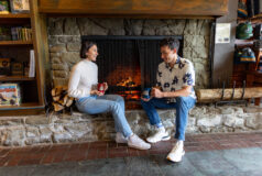 Two people sit by a stone fireplace, smiling and holding mugs in a cozy, rustic room in Kalaloch Main Lodge.