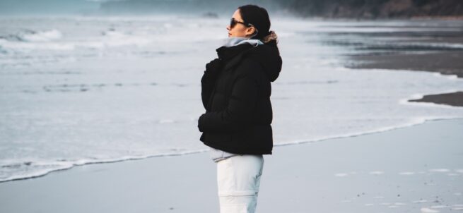 Person in a black jacket and white pants standing on a beach in Olympic National Park, looking out at the ocean.