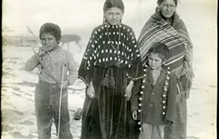 Four Indigenous people, a woman and three children, stand on snowy ground wearing traditional clothing.