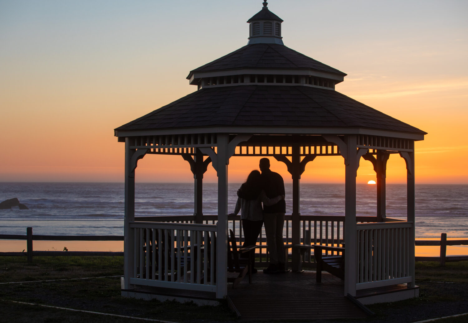 Two people embrace in a gazebo, watching the sunset over the ocean.