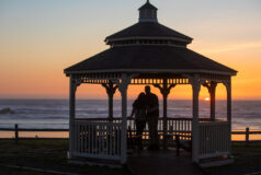 Two people embrace in a gazebo, watching the sunset over the ocean.
