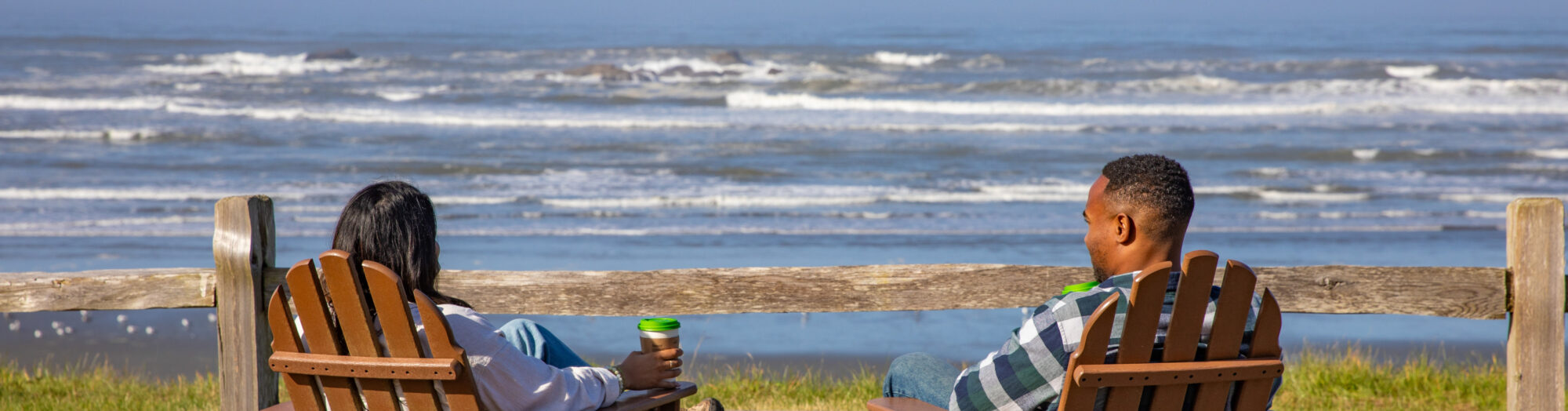 Two people sit in wooden chairs facing the ocean, enjoying a sunny day by the beach.