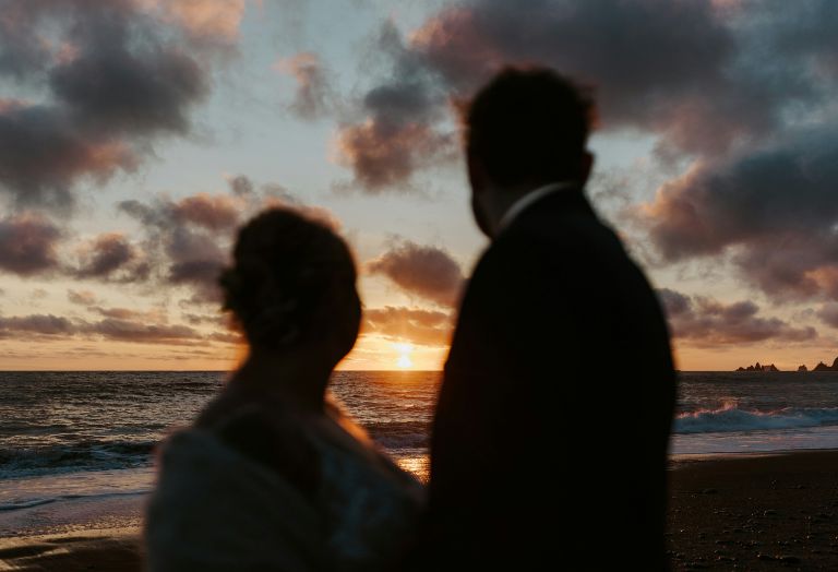 A couple in silhouette stands on a beach, watching the sunset over the ocean with clouds in the sky.