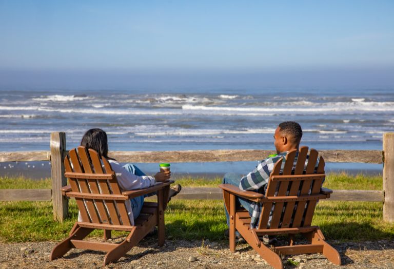 Two people sit in wooden chairs facing the ocean, enjoying a sunny day by the shore.