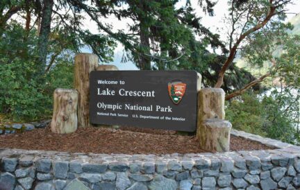 A sign for Lake Crescent at Olympic National Park, surrounded by trees and a stone wall.